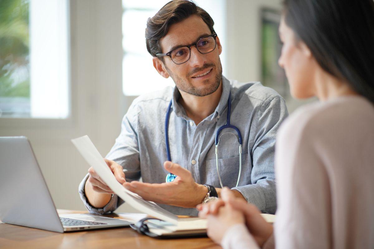 Doctor with a stethoscope discusses paperwork with a patient at a desk, with a laptop and notebook in view.