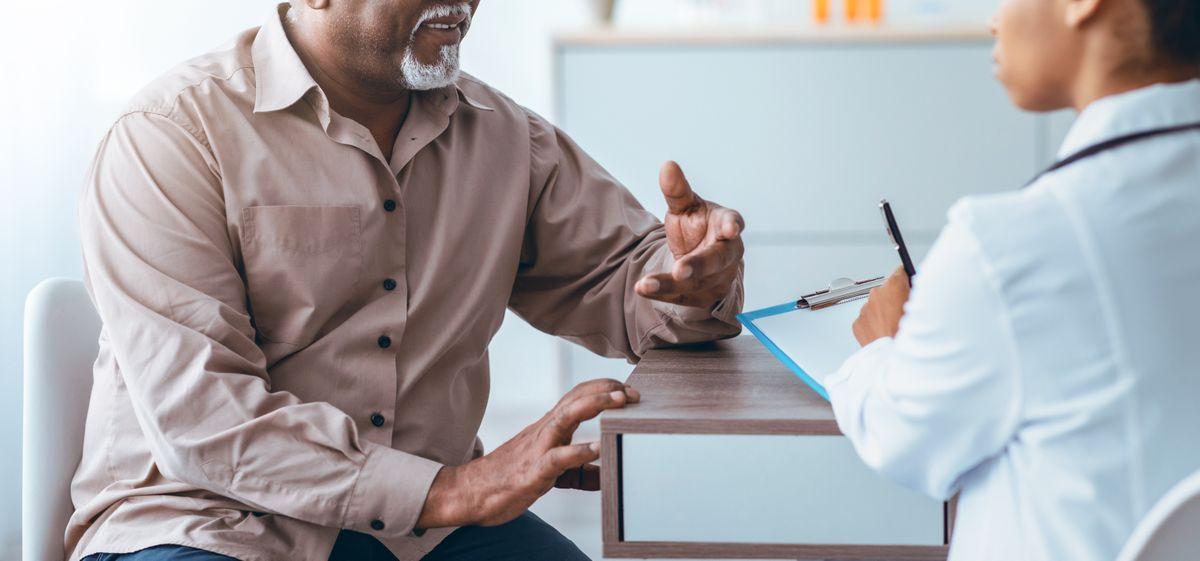 A man in a beige shirt gestures while speaking to a doctor taking notes on a clipboard, in a medical office setting.