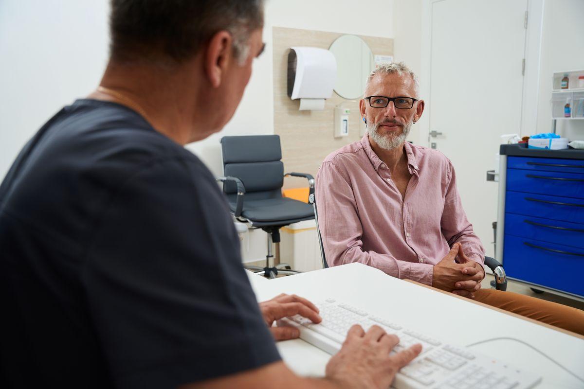 Doctor typing at a desk in a medical office while a male patient in glasses and a pink shirt sits attentively across from him.