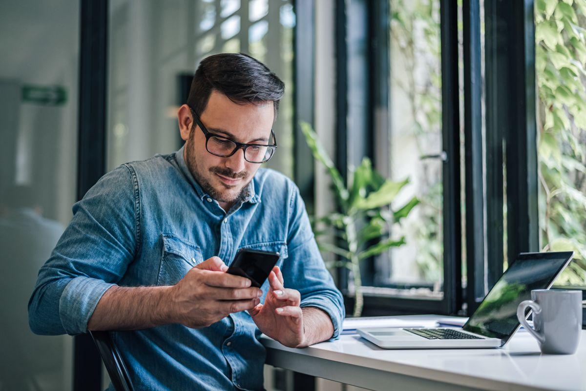 A man in glasses and a denim shirt uses a smartphone while sitting at a desk with a laptop, near a window with plants.